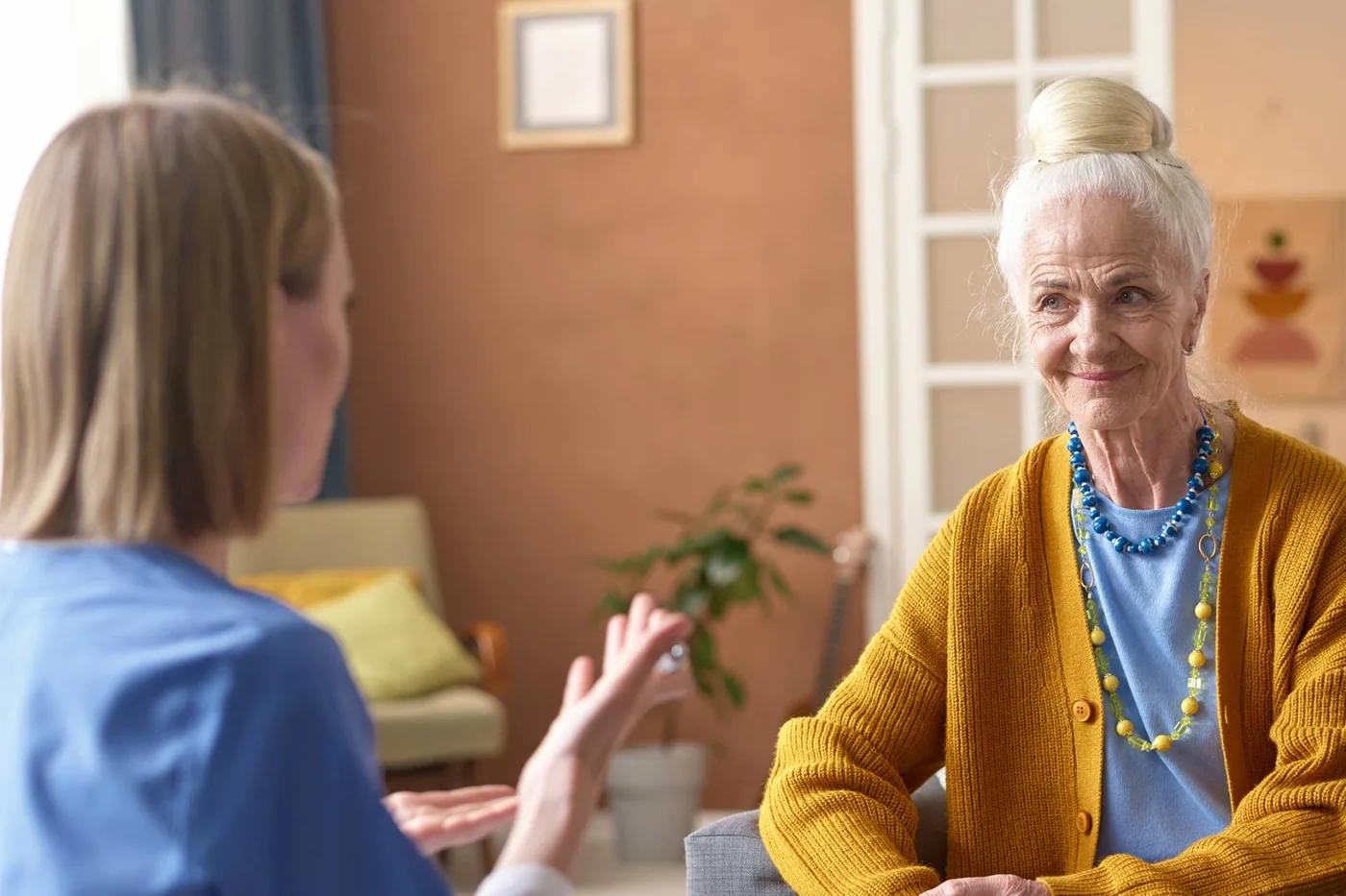 Hörakustikerin berät eine ältere Dame in gelber Strickjacke in deren Wohnzimmer